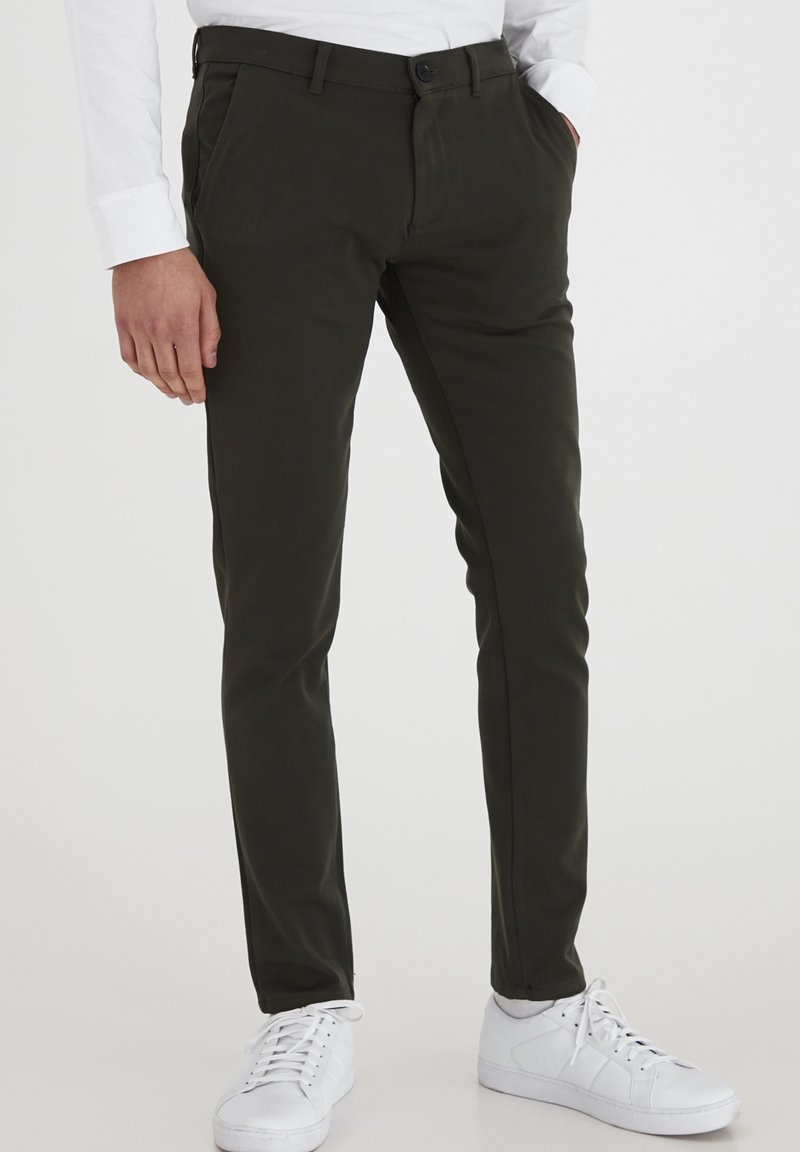 Dunkelgrüne Hose mit Slim Fit, aus glattem Stoff gefertigt, mit seitlichen Taschen und Knopfverschluss, kombiniert mit weißen Sneakers.