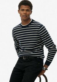 Marineblauwe gestreepte trui met witte horizontale strepen, geribbelde halslijn en manchetten, gecombineerd met zwarte broek en een horloge om de pols.