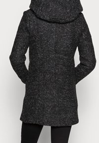 Cappotto grigio scuro in misto lana con un grande cappuccio morbido, tessuto texturizzato, silhouette aderente e taglio a lunghezza media. La vista posteriore mostra i dettagli delle cuciture.