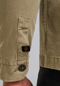 Khaki plátená bunda s rukávom s gombíkovým zapínaním a šitými detailmi. Obsahuje čiernu nášivku s logom "PME Legend". Hladká textúra.