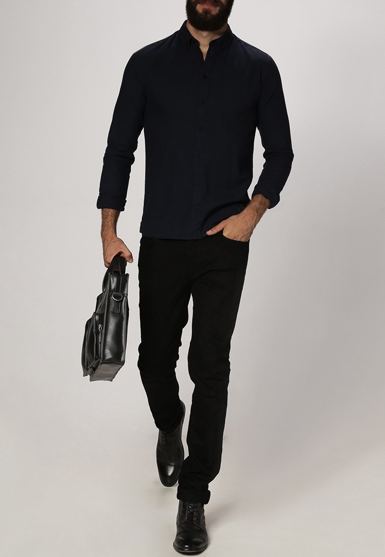 Donkerblauwe overhemdd, zwarte broek en zwarte leren schoenen. Houdt een zwarte leren tas vast. Slim fit, lange mouwen en kraagontwerp.