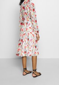 Blumenkleid mit weißem Grund und roten, pinken sowie gelben Blumen, langen Ärmeln, gestuftem Rock und kombiniert mit braunen Riemchensandalen.