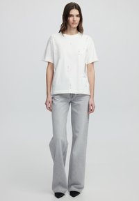 T-shirt blanche à manches courtes avec des clous, associée à un jean évasé gris clair. Les deux pièces ont une coupe décontractée et un design minimaliste.