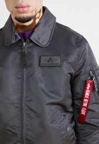 Black nylon bomber jacket featuring a high collar, zip-up front, and an "Alpha Industries" logo patch. Includes a red "Remove Before Flight" tag.