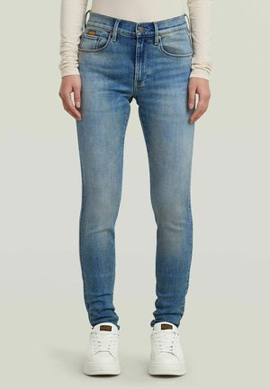 Vue de face d'une personne portant un jean slim bleu clair, un haut beige à manches longues et des baskets blanches, debout devant un fond pâle.
