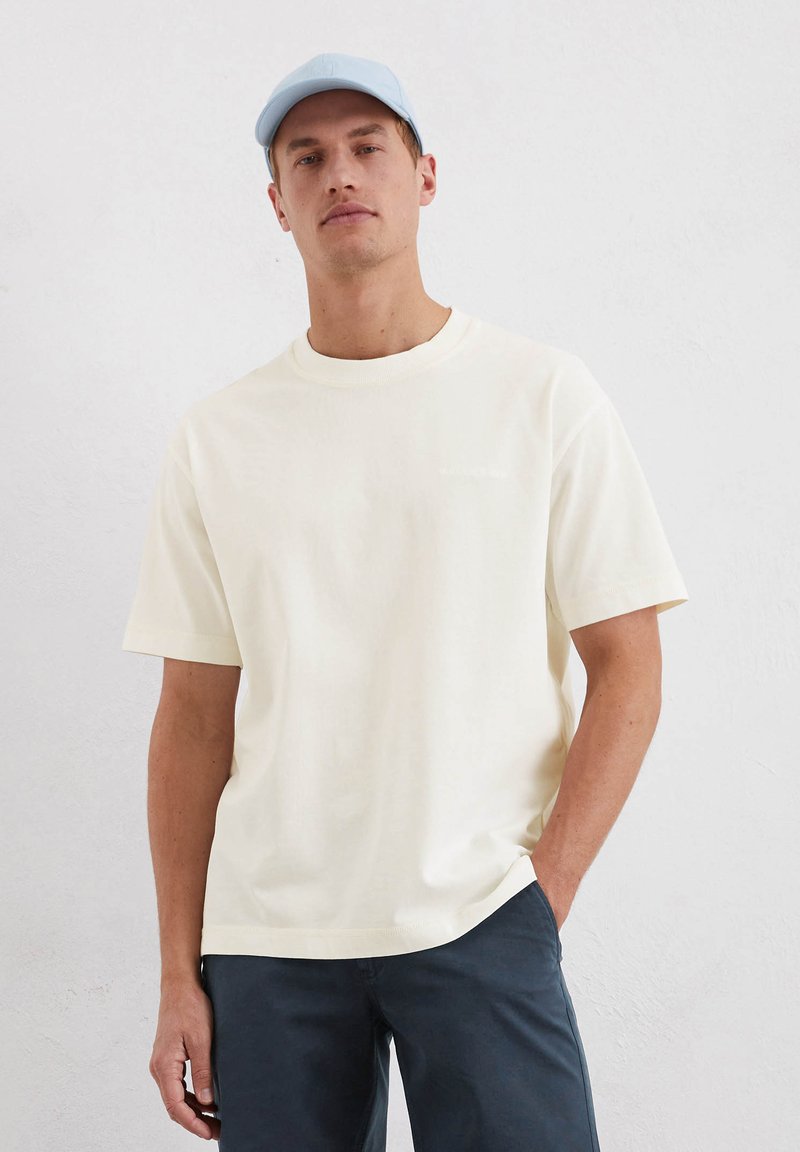 Cream short-sleeve t-shirt with a round neckline and relaxed fit. Paired with dark pants. Minimalist design with subtle branding.
