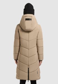 Beige vadderad jacka med huva, med fiskbenssöm-design, framdragkedja och en ribbad svart stickad mössa.