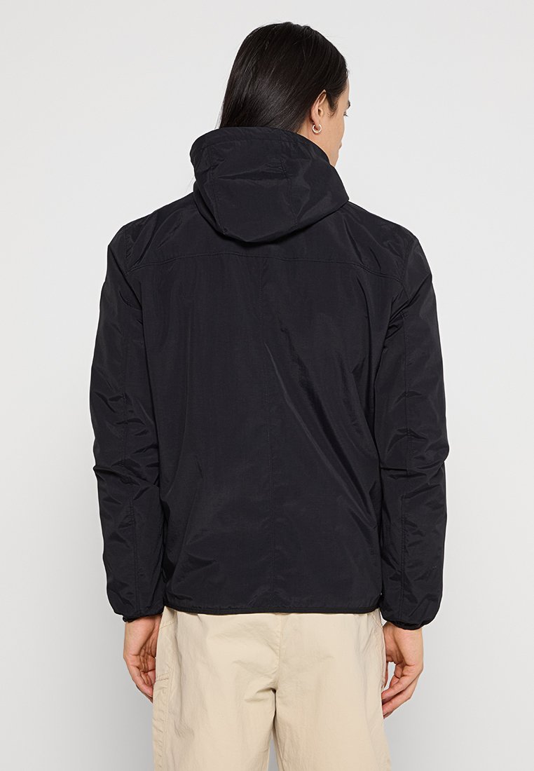 Giacca nera con cappuccio, finitura liscia e opaca. Presenta una vestibilità rilassata e cuciture sulle spalle distintive. Nessun hardware o motivo visibile.