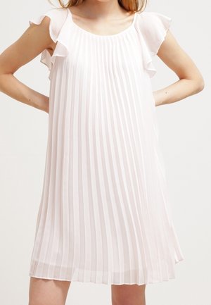 Femme portant une robe blanche plissée sans manches avec des détails à volants aux épaules, debout, les mains sur les hanches.
