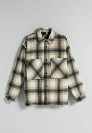 Homme portant une veste à carreaux beige à boutons, un pantalon cargo beige clair et des baskets noires, blanches et marron, debout sur un fond gris.