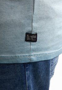 Lyseblå t-shirt med en glat tekstur, udstyret med et sort stofmærke med hvid tekst, parret med mørkeblå denim jeans.
