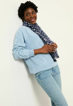 Light blue sweater with textured knit, paired with a patterned navy scarf. Slim-fit denim jeans complete the outfit.