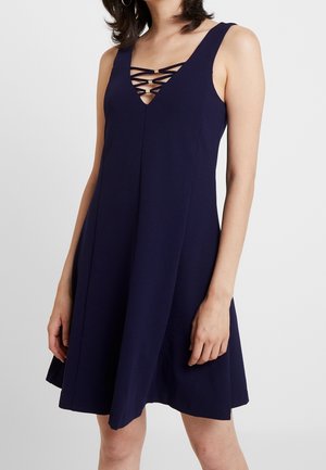 Robe bleu marine sans manches avec un décolleté en V et un détail de bretelles croisées, portée par une femme aux cheveux foncés et boucles d'oreilles anneaux.
