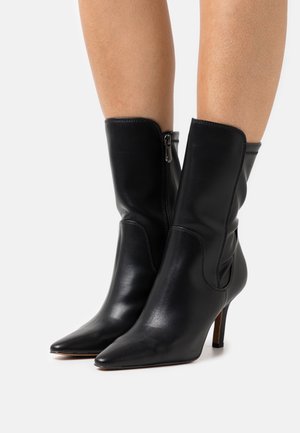 Botas de tobillo de cuero negro con punta afilada, cierre de cremallera en el costado y un tacón alto y delgado. Textura suave y diseño ajustado.