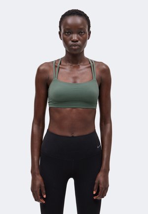 Kvinde med mørk hud iført en grøn sports-bh og sorte højttaljede leggings, der står mod en ensfarvet lys baggrund.