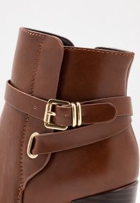 Hnědá kotníková kožená bota s hladkou texturou, zdobená zlatou přezkou a šitými detaily podél okrajů.