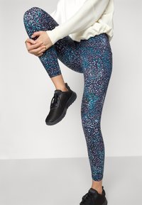 Sötétkék, türkiz és fehér színű, többszínű pettyes mintázattal ellátott leggings, rugalmas anyagból készült, fekete sportcipővel párosítva.