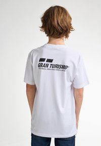 Bílá bavlněná tričko s krátkým rukávem. Na zadní straně se nachází "GRAN TURISMO" v černém tučném písmu a pod tím "SKUTEČNÝ ŘÍDICÍ SIMULÁTOR".