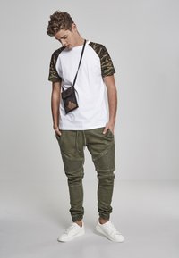White T-shirt with camouflage sleeves, olive green joggers with textured knee accents, and white sneakers. Small black pouch worn crossbody.