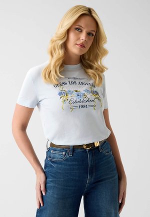 Femme blonde aux cheveux ondulés portant un t-shirt graphique floral bleu clair "Guess Los Angeles" et un jean taille haute bleu avec une ceinture marron.