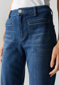 Blauwe denim jeans hebben een middelhoge taille, voorzakken en achterzakken met gestikte details. De stof heeft een gladde textuur.