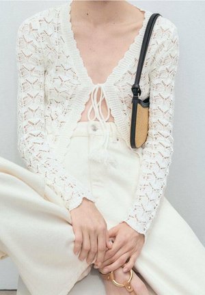 Personne portant un haut court à manches longues en crochet blanc, un pantalon crème taille haute, tenant un sac à bandoulière tissé beige et noir.