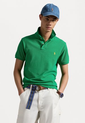 Groene poloshirt met kraag en korte mouwen, gecombineerd met witte broek en een bruine riem met blauwe en gele strepen. Denim pet.