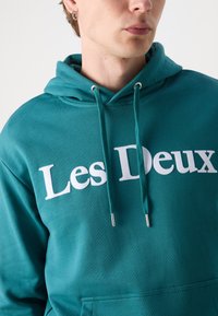 Teal hoodie made of soft fabric, featuring a large white "Les Deux" logo, drawstrings with silver tips, and a front kangaroo pocket.