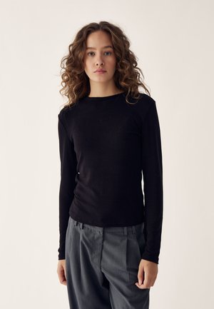 Camicia nera a maniche lunghe in tessuto morbido con scollo rotondo, abbinata a pantaloni grigi plissettati con vestibilità comoda. Texture liscia, design minimalista.