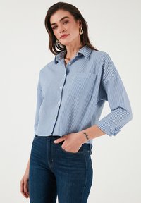 Blue and white striped button-up shirt with a collar, two chest pockets, and rolled three-quarter sleeves, paired with dark blue jeans.