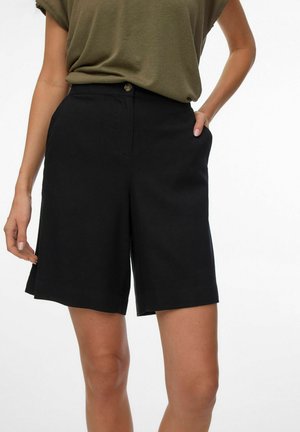 Donna che indossa pantaloncini neri ad altezza vita fino al ginocchio con bottone e tasche laterali, abbinati a una maglietta verde oliva infilata dentro.