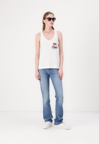 Witte katoenen tanktop met een ronde hals en een kleurrijke logo patch, gecombineerd met lichtblauwe spijkerbroeken en witte sneakers. Zwarte zonnebril op.