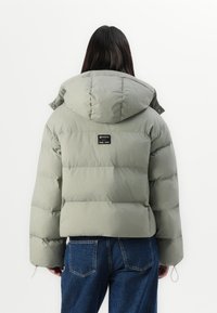 Person med langt mørkt hår iført en lysegrå pufferjakke med hætte og blå denimjeans, set fra ryggen mod en hvid baggrund.