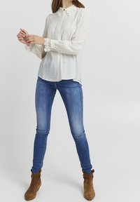 Frau trägt eine Bluse mit weißer Spitzenkragen, enge blaue Jeans und braune Wildleder-Stiefeletten, stehend vor einem schlichten Hintergrund.