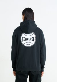 Sudadera negra con un gran logo circular blanco de "Converse" en la parte trasera, con costuras de acento. Material suave de mezcla de algodón, ajuste casual.