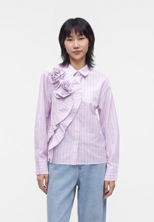 Jeune femme aux cheveux noirs porte un chemisier rayé violet clair avec des détails floraux à volants et un jean bleu clair sur un fond uni.