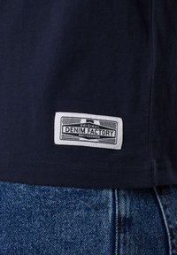 Navy-blaues T-Shirt mit einem weißen Etikett, auf dem "ORIGINAL DENIM FACTORY" aufgenäht ist, kombiniert mit blauen Jeans.