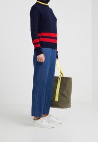 Personne portant un pull marine à rayures rouges, un pantalon large bleu, des baskets blanches, tenant un sac fourre-tout olive avec des anses jaunes.