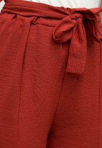 Pantaloni color ruggine con cintura annodata, tessuto testurizzato e tasche laterali. Presentano delicate pieghe e un design a vestibilità rilassata.