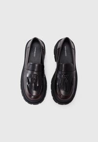 Pantofi loafers din piele neagră cu vârful rotunjit, având ciucuri în partea din față și o talpă robustă neagră pentru o aderență suplimentară.