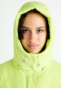 Lime green puffer jacket with a high hood, snap buttons, and adjustable drawstrings. Quilted texture adds visual interest.