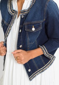 Džínová bunda v tmavě modré barvě s třepenými okraji, zlatým prošíváním a stříbrnými knoflíky. Obsahuje dvě přední kapsy a zkrácený střih.