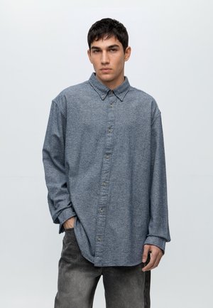 Junger Mann steht mit der Hand in der Tasche, trägt ein lockeres graues Hemd mit Knöpfen und ausgewaschene schwarze Jeans vor einem schlichten weißen Hintergrund.