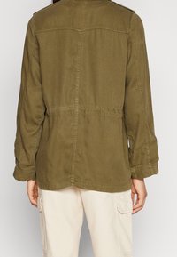 Bawełniana kurtka w oliwkowym zielonym kolorze o luźnym kroju, z widocznym przeszyciem, zapinanymi na guziki pagonami na ramionach oraz elastycznym detailem w pasie.