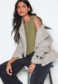 Beige trenchcoat med store sorte knapper, lag på lag over en grøn top. Mørke, løstsiddende bukser. Glat stof, afslappet pasform, stilfuld skræddersyning.