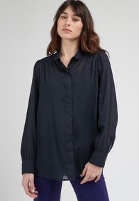 Blusa blu navy, tessuto leggero, chiusura con bottoni davanti, maniche lunghe con polsini arricciati, taglio dritto, leggero luccichio.