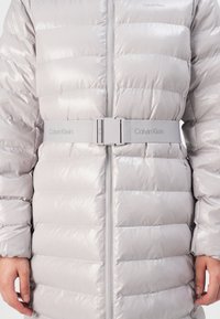 Lys grå pufferjakke med horisontal quiltet design, glidelåsfront og matchende belte i midjen, med Calvin Klein-merke på beltet.