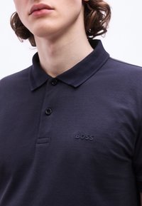 Νεαρό άτομο που φοράει σκούρο μπλε πόλο μπλουζάκι με γιακά και δύο κουμπιά, με ένα μικρό λογότυπο "BOSS" στο στήθος.