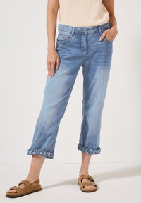 Lichtblauwe denim broek met een ontspannen pasvorm, opgerolde broekspijpen met een patroonrand en voorzakken. Gepaard met bruine sandalen.