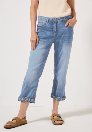 Lichtblauwe denim broek met een ontspannen pasvorm, opgerolde broekspijpen met een patroonrand en voorzakken. Gepaard met bruine sandalen.